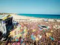 Multitud vibrante en el Arenal Sound Valencia, lanzando polvo de colores bajo el sol. El mar azul y la playa de fondo crean un escenario festivo.