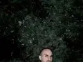 Un hombre calvo, con mirada intensa, posa frente a un fondo de follaje oscuro. Podría ser un sicario rapero, listo para soltar rimas letales.
