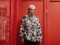 El cantante Kaze, con gorra blanca y chaqueta polar con grafitis, posa en un pasillo rojo vibrante.