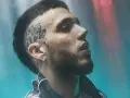 Un joven **hard cantante** con tatuaje en la cabeza y barba, mirando hacia arriba con luces de neón de fondo.