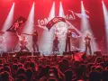 Una banda toca en un escenario iluminado con luces rojas, ondeando banderas piratas. El público, con trece roeis, anima la actuación.