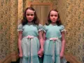 Dos niñas idénticas, vestidas con trajes azules, se toman de la mano en un pasillo con papel tapiz floral. Son las gemelas de "Bso El Resplandor".