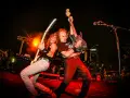 Dos guitarristas de las ruedas grupo musical tocan con pasión en un escenario iluminado por luces rojas y naranjas.