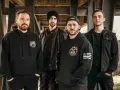 Cuatro hombres con sudaderas y chaquetas negras posan bajo una estructura metálica. Sus miradas intensas parecen **blaze out** con determinación.