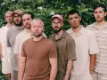 Los miembros de Oques Grasses posan juntos, con ropa de tonos tierra y un fondo de vegetación frondosa.