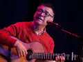 Mario Díaz, con gafas y un suéter rojo, toca la guitarra y canta frente a un micrófono en un escenario con luces tenues.