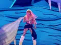Karol G, con su cabello rosa vibrante, canta en el escenario con un traje brillante y botas peludas. Un pez inflable gigante la acompaña en esta presentación.
