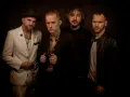 El grupo de embusteros posa con miradas intensas. Uno lleva sombrero y traje blanco, otro camisa de leopardo, y los otros dos, chaquetas oscuras.