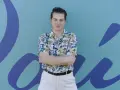 Joven con camisa tropical y pantalones claros, brazos cruzados, frente a un mural azul con formas orgánicas. Parece un miembro del ortiga grupo.