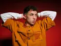Joven con las manos detrás de la cabeza, vistiendo una chaqueta naranja y una camiseta blanca. Parece un pole cantante listo para actuar.