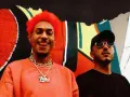 Dj Ballesteros y su colega posan frente a un mural vibrante. El primero, con cabello rojo y un collar dorado, sonríe con la lengua afuera.