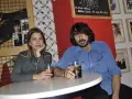Pareja en un bar con decoración rockera. Ella, con camisa de cuadros, apoya la barbilla en la mano. Él, con camisa azul, sostiene un vaso. La furia trinidad de la música inspira el ambiente.