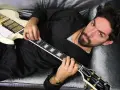 El cantante Siloe, con barba y cabello oscuro, toca una guitarra eléctrica blanca con detalles dorados, recostado en un sofá de terciopelo gris.