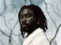 Retrato de un rapero español con rastas y un pendiente plateado, vistiendo una chaqueta clara.