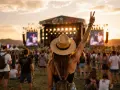 Una chica con sombrero levanta la mano en señal de paz en un festival al atardecer. La multitud disfruta de la música bajo el cielo dorado.
