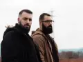 Dos hombres con barba posan con seriedad. Uno viste chaqueta negra, el otro marrón. Parecen listos para un rap de "falsa alarma".