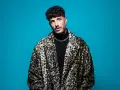 Wade DJ, con su abrigo de leopardo y cadena plateada, posa con confianza ante un fondo azul vibrante.