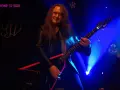 Guitarrista con pelo largo y chaqueta de cuero, tocando una guitarra eléctrica negra con detalles rojos. El escenario está iluminado con luces azules, creando un ambiente de rock.