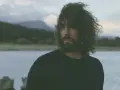 Un hombre con cabello rizado y barba mira hacia el horizonte, entre el miedo y el paraíso de un paisaje acuático y montañoso.