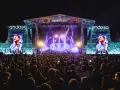 Gran concierto en el Jardín de las Delicias. Multitud vibrante ante el escenario iluminado, con pantallas gigantes mostrando al artista.