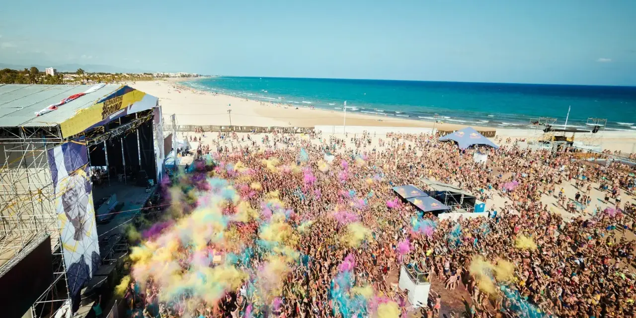 Multitud vibrante en el Arenal Sound Valencia, lanzando polvo de colores bajo el sol. El mar azul y la playa de fondo crean un escenario festivo.