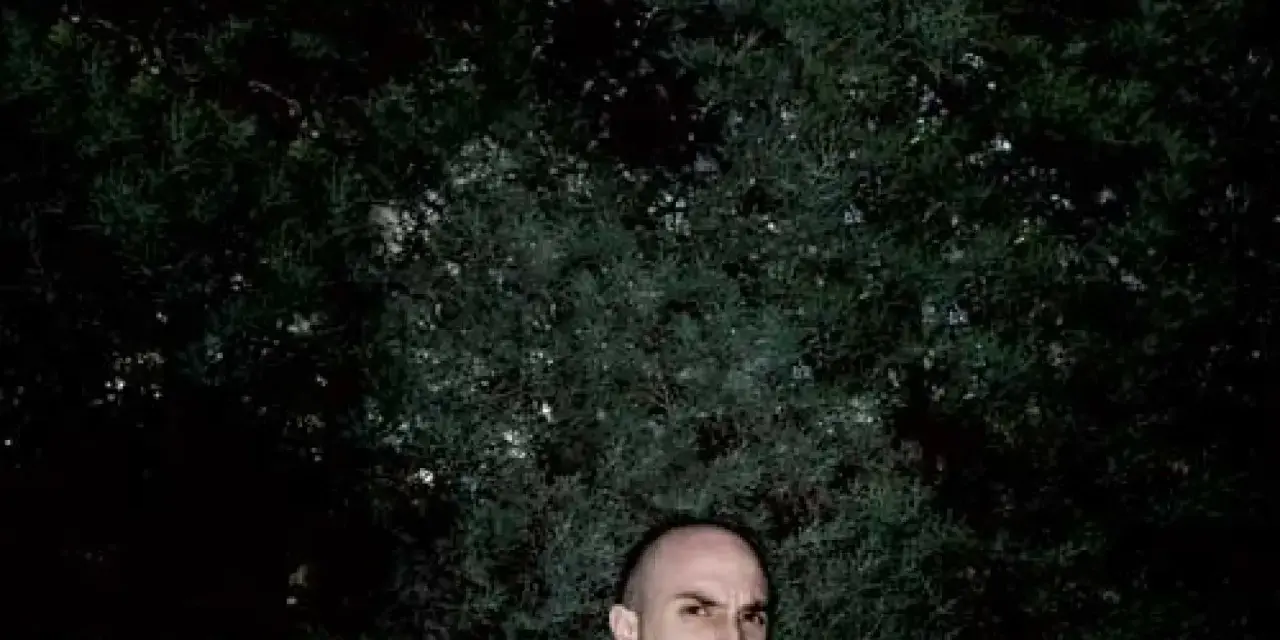 Un hombre calvo, con mirada intensa, posa frente a un fondo de follaje oscuro. Podría ser un sicario rapero, listo para soltar rimas letales.