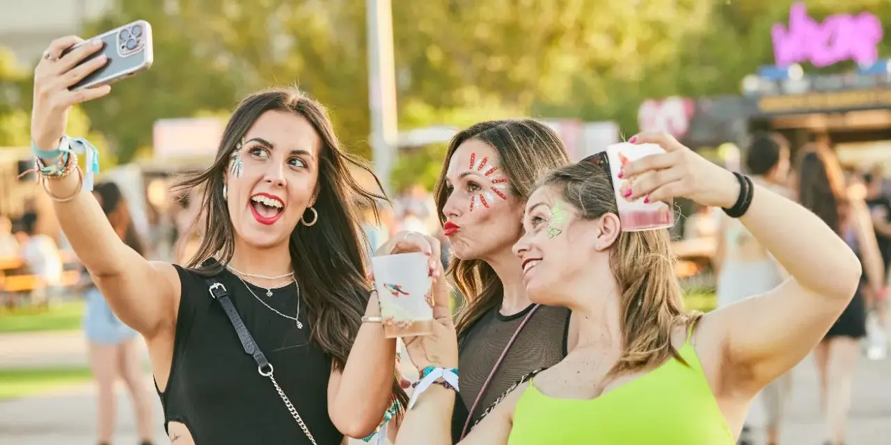Tres amigas con maquillaje festivo y bebidas en un festival de verano.