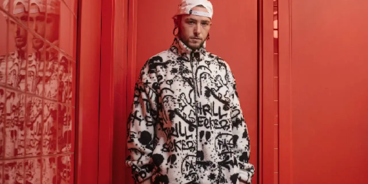 El cantante Kaze, con gorra blanca y chaqueta polar con grafitis, posa en un pasillo rojo vibrante.