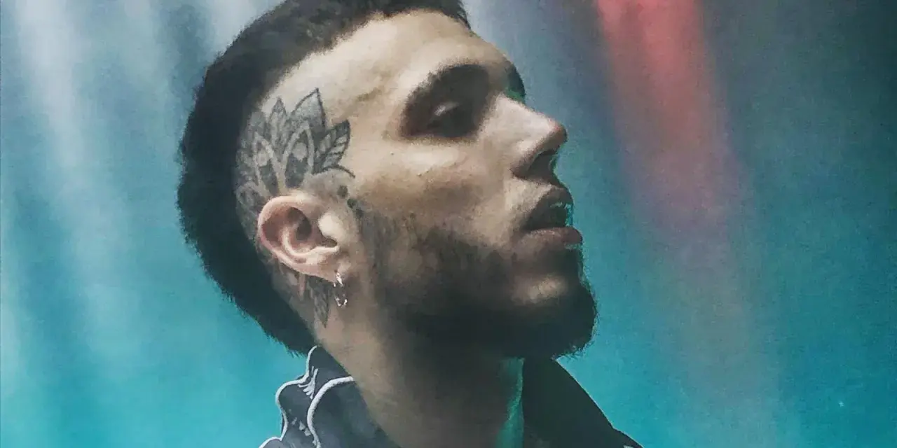 Un joven **hard cantante** con tatuaje en la cabeza y barba, mirando hacia arriba con luces de neón de fondo.