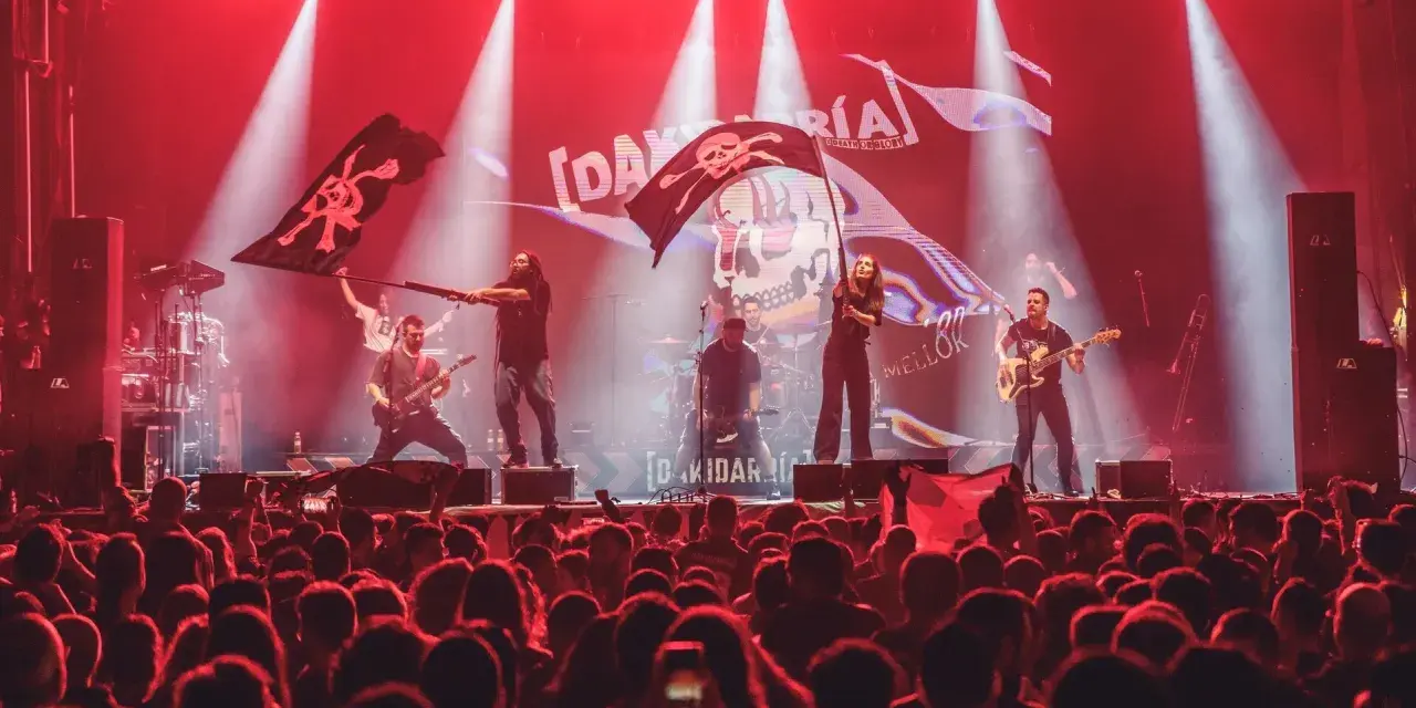 Una banda toca en un escenario iluminado con luces rojas, ondeando banderas piratas. El público, con trece roeis, anima la actuación.