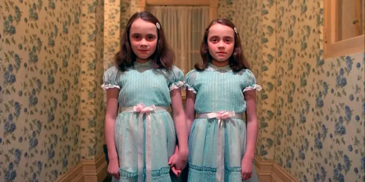 Dos niñas idénticas, vestidas con trajes azules, se toman de la mano en un pasillo con papel tapiz floral. Son las gemelas de "Bso El Resplandor".