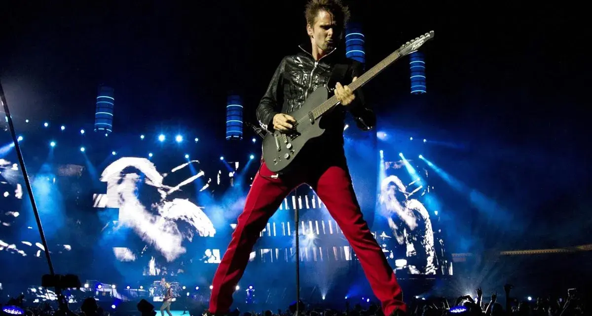 Matt Bellamy de Muse España, con pantalones rojos y chaqueta negra, toca la guitarra en un escenario iluminado por luces azules y pantallas gigantes.