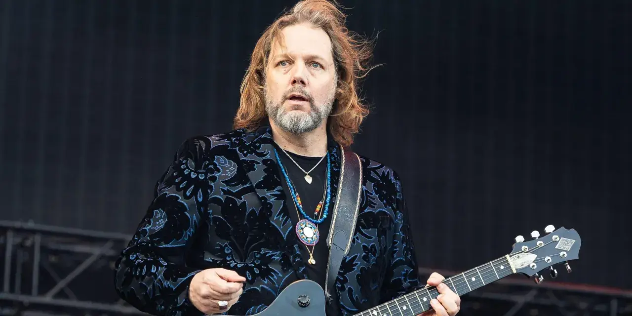 Rich Robinson, guitarrista con cabello largo y barba, luce un saco negro con estampado floral azul y varias cadenas.