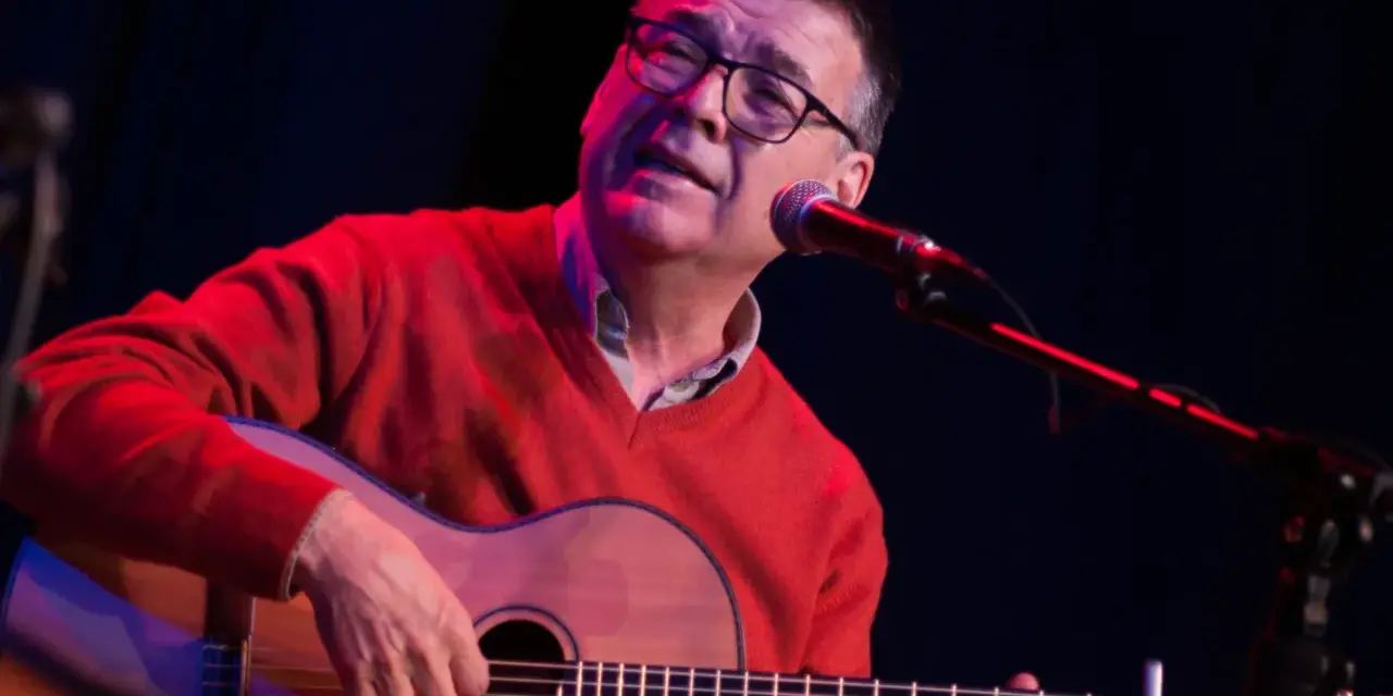 Mario Díaz, con gafas y un suéter rojo, toca la guitarra y canta frente a un micrófono en un escenario con luces tenues.