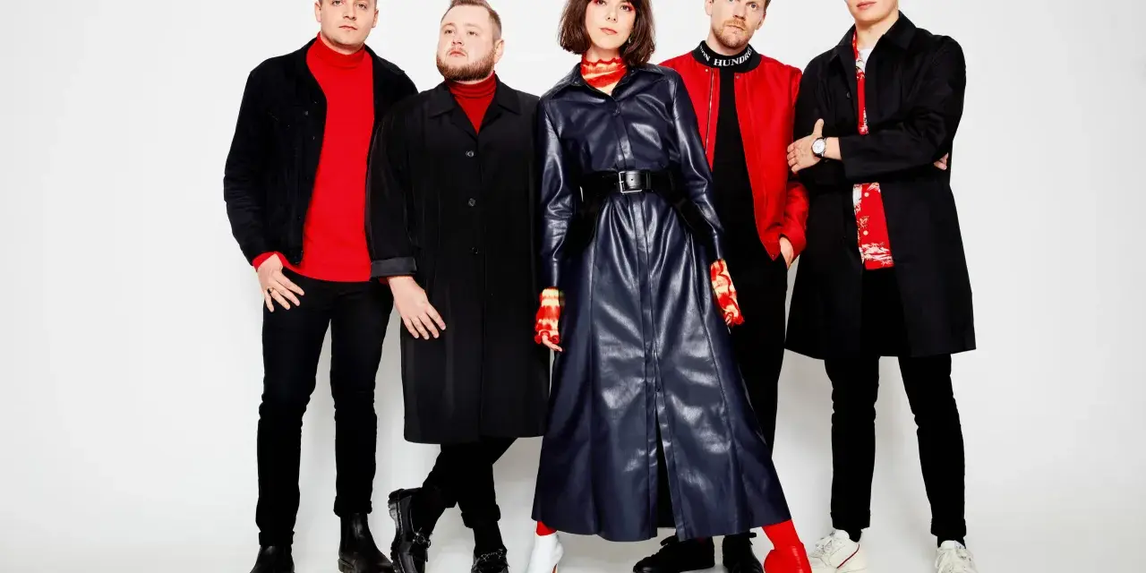 Cinco personas posan con ropa negra y roja. El cartel "San San" podría ser el nombre de la banda.