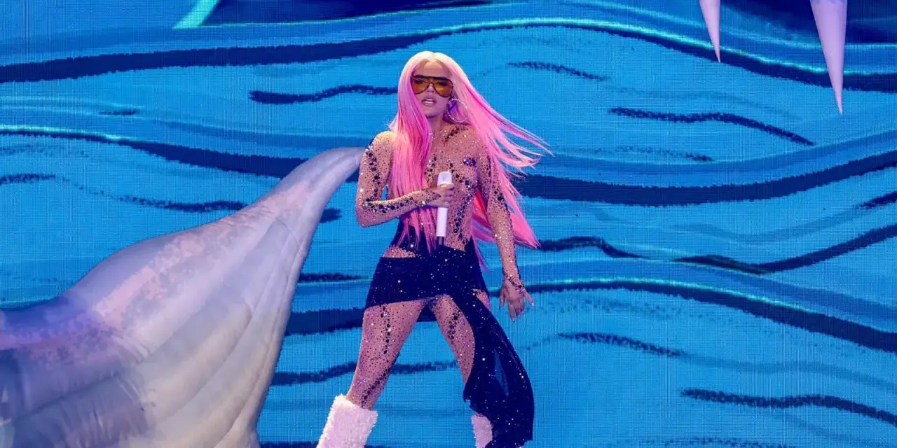 Karol G, con su cabello rosa vibrante, canta en el escenario con un traje brillante y botas peludas. Un pez inflable gigante la acompaña en esta presentación.