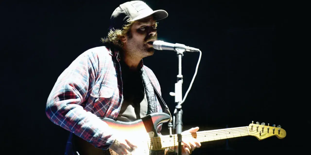 Neil Halstead, con gorra y camisa a cuadros, toca la guitarra y canta en un concierto.