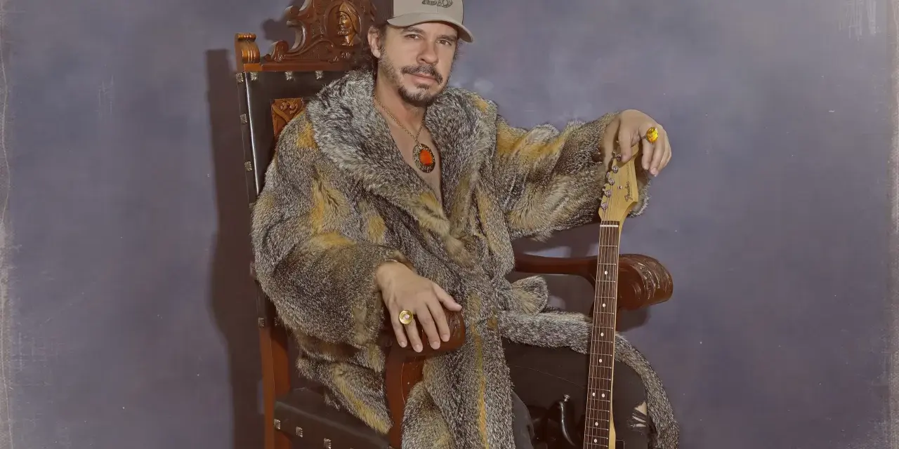 Luis Brea, con gorra y abrigo de piel, sentado en un trono con una guitarra.