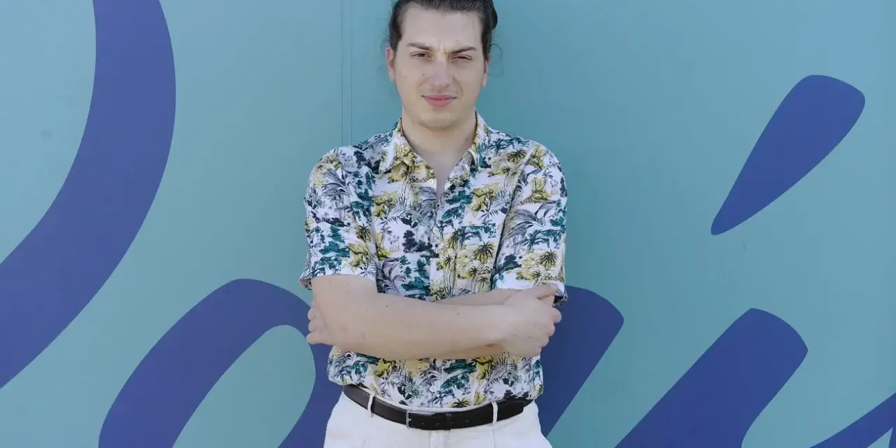 Joven con camisa tropical y pantalones claros, brazos cruzados, frente a un mural azul con formas orgánicas. Parece un miembro del ortiga grupo.