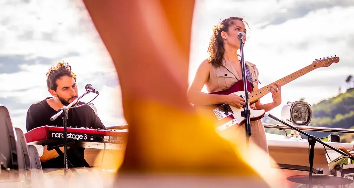 Maria y Feu, una banda en vivo, tocan en un escenario al aire libre. Ella canta y toca la guitarra, él en los teclados.