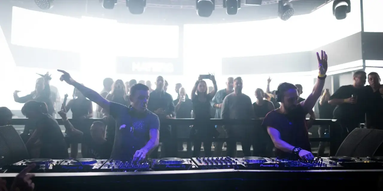 Dos DJs actuando en un club con público al fondo.