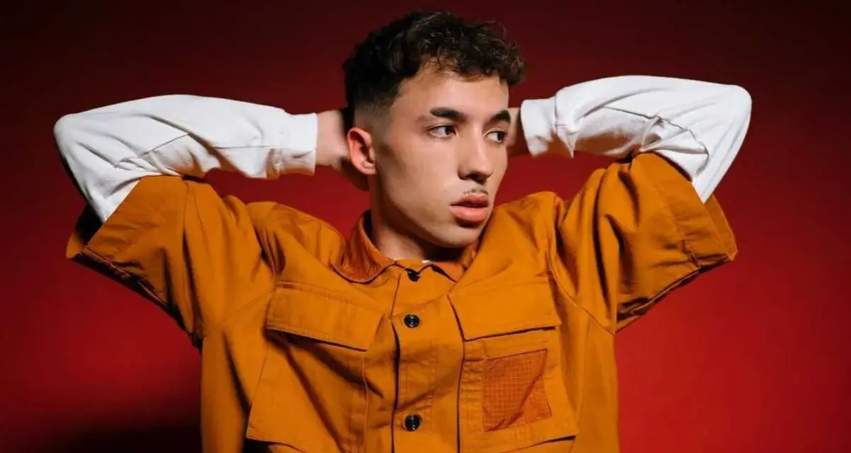 Joven con las manos detrás de la cabeza, vistiendo una chaqueta naranja y una camiseta blanca. Parece un pole cantante listo para actuar.