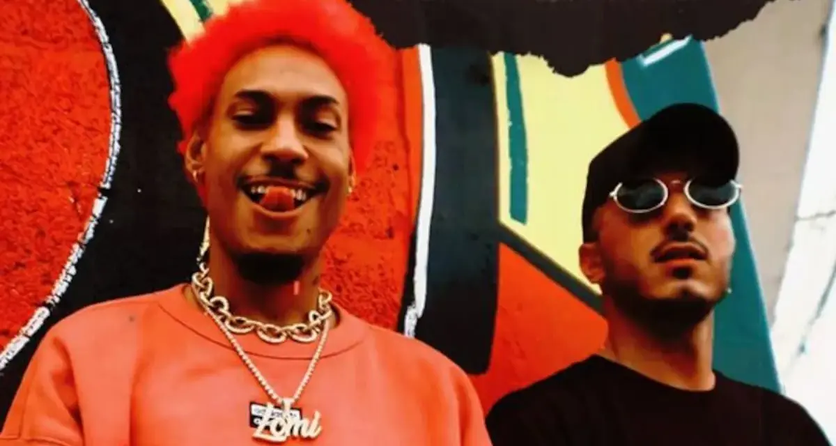 Dj Ballesteros y su colega posan frente a un mural vibrante. El primero, con cabello rojo y un collar dorado, sonríe con la lengua afuera.