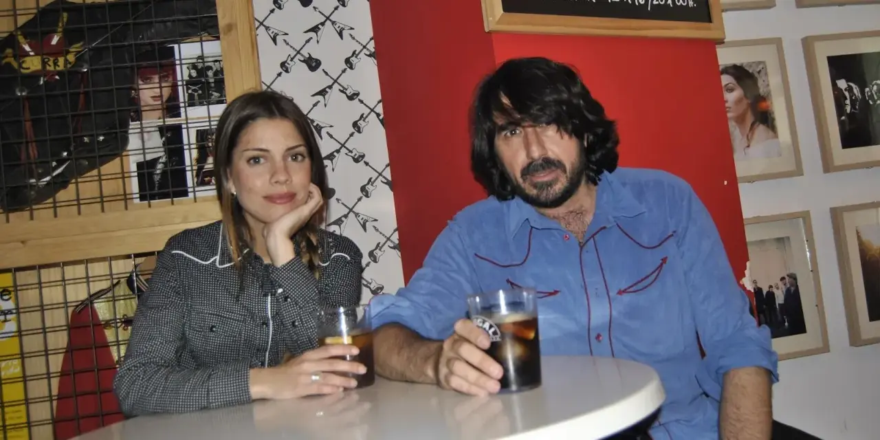 Pareja en un bar con decoración rockera. Ella, con camisa de cuadros, apoya la barbilla en la mano. Él, con camisa azul, sostiene un vaso. La furia trinidad de la música inspira el ambiente.