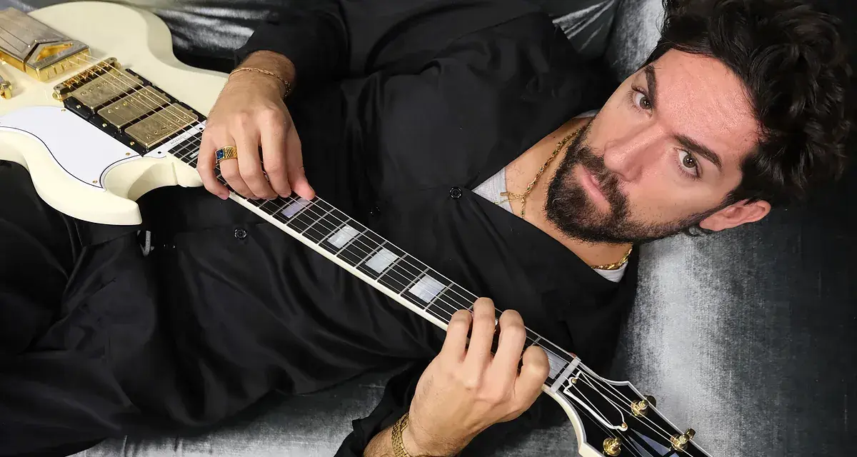El cantante Siloe, con barba y cabello oscuro, toca una guitarra eléctrica blanca con detalles dorados, recostado en un sofá de terciopelo gris.