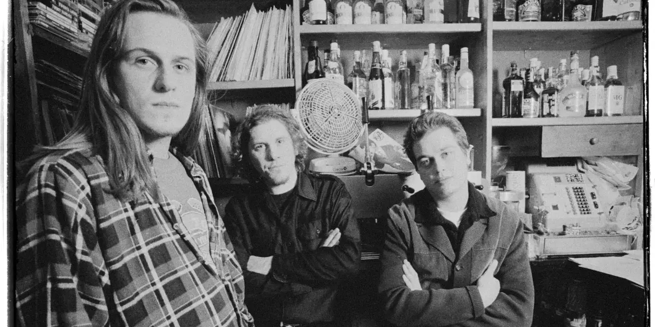 Tres hombres posan en blanco y negro, con estanterías de discos y botellas detrás.