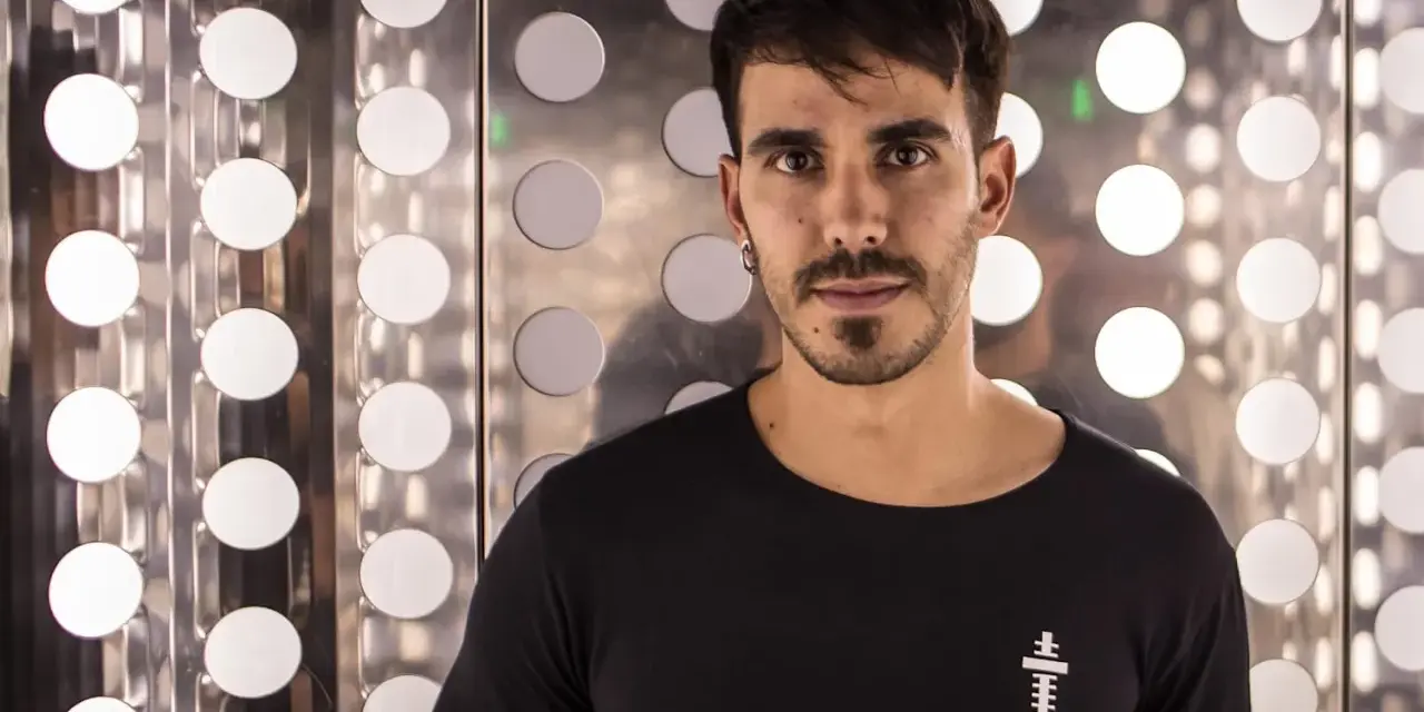 Raúl Pacheco, DJ, posa con una camiseta negra frente a un fondo metálico con círculos blancos brillantes.
