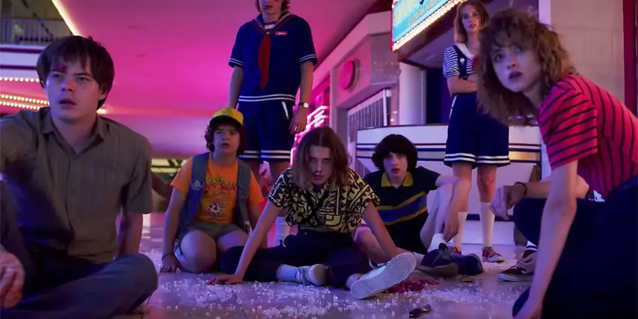 Los personajes de Stranger Things, con Eleven al frente, sentados en el suelo de un centro comercial, evocando las icónicas canciones de la serie.