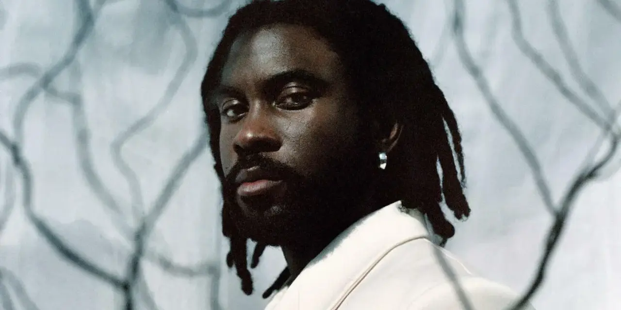 Retrato de un rapero español con rastas y un pendiente plateado, vistiendo una chaqueta clara.