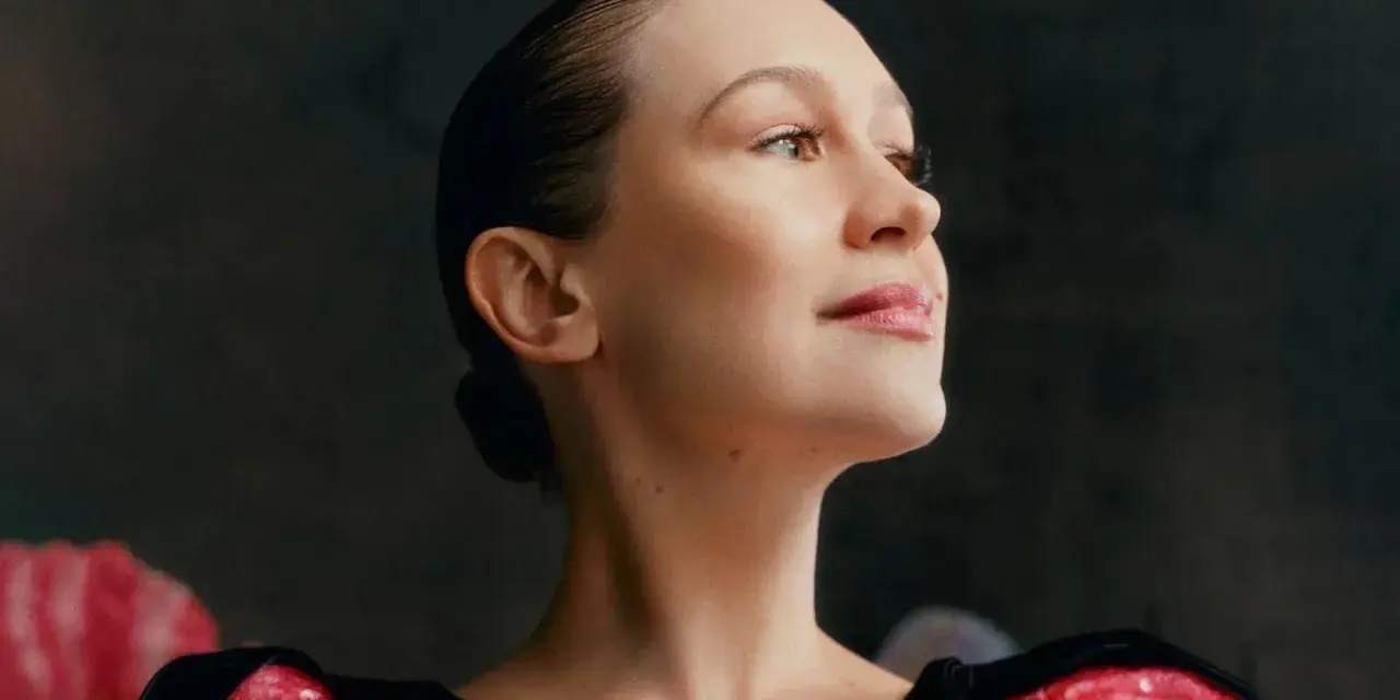 Joanna Newsom, con su cabello recogido y un vestido negro con detalles brillantes, mira hacia arriba con una expresión serena.